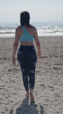 Genç, uzun saçlı esmer, beyaz bir kadın güneşli bir günde kumsalda geriye doğru yürüyen ellerini kaldırarak hayranları FullHD dikey