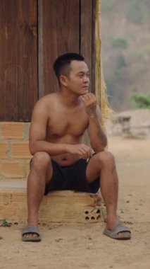 Çıplak gövdeli genç Vietnamlı bir adam evinin kapısında dinlenirken sigara içiyor - FullHD Dikey