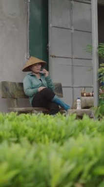 Olgun Asyalı kadın çiftçi ahşap bir bankta oturmuş Vietnam 'da akıllı telefonuyla konuşuyor.