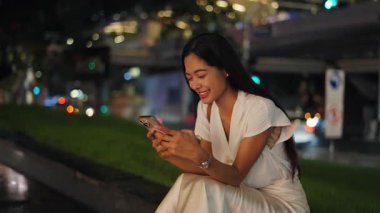 Şehrin ana caddesinin yanında oturan Asyalı genç kadın sıcak bir gecede akıllı telefonuyla mesajlar gönderiyor - 4K Yatay