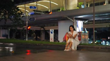 Asyalı kız çimlerin üzerinde oturmuş Bangkok 'taki büyük caddenin yanındaki akıllı telefon ekranına bakıyor - 4K Yatay