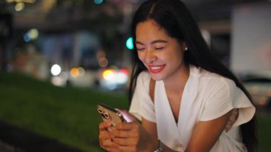 Geceleri şehirde oturan akıllı telefonuyla fotoğraflara bakan genç bir kadının yakın çekim yüzü ve elleri - 4K Yatay