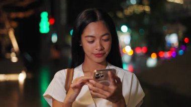 Gece akıllı telefonunun ekranına dokunurken kameraya gülümseyen genç bir kadının yüzü ve elleri - 4K Yatay