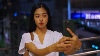 Sıska, genç Asyalı kız akıllı telefonunun önünde Tayland 'da gece vakti büyük şehirde selfie çekmek için el kol hareketi yapıyor.