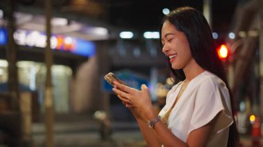 Uzun düz saçlı Asyalı kız gece Bangkok 'taki büyük caddede akıllı telefonuyla video görüşmesi yapıyor. 4K Yatay.