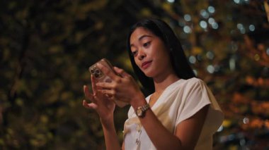 Yaz gecesi bileğinde saati olan Asyalı esmer kız akıllı telefon ekranına dokunuyor - 4K Yatay