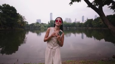 Kalp şeklinde güneş gözlüğü takan genç bir kadın parkta akıllı telefon kullanıyor - 4K Yatay