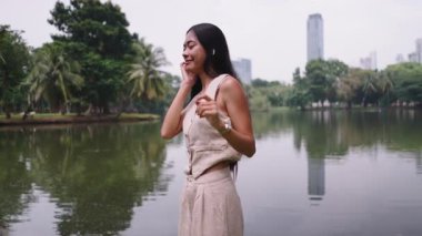 Genç Asyalı iş kadını kablosuz kulaklıklarla müzik dinliyor ve şehir parkında göl kenarında dans ediyor - 4K Yatay