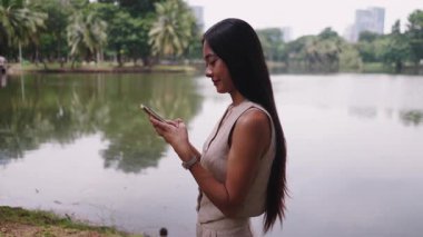 Şehir parkında gölün yanında akıllı telefon kullanan genç bir iş kadını - 4K Yatay