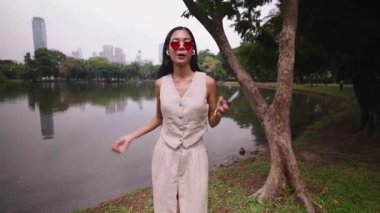 Kalp şeklinde güneş gözlüğü takan genç bir kadın parkta dans ediyor ve müzik dinliyor - 4K Yatay