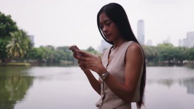 Şehir parkında gölün yanında akıllı telefon kullanan Asyalı genç bir iş kadını - 4K Yatay