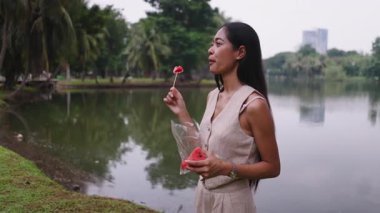 Genç bir kadın, sakin bir gölün kenarındaki çubukta ferahlatıcı bir karpuz diliminin tadını çıkarıyor - 4K Yatay