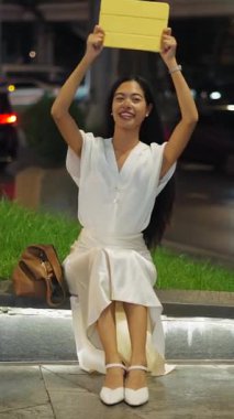 Bangkok 'ta geceleri elinde tablet tutarken kameraya bakıp gülümseyen Asyalı genç bir kadın.