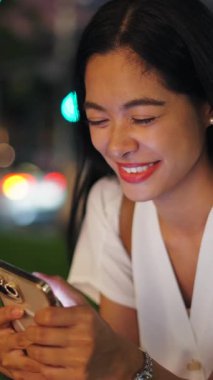 Geceleri akıllı telefonuyla fotoğraflara bakan genç bir kadının yüzü ve elleri şehirde dikey bir şekilde oturuyor.