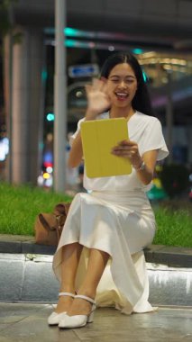 Genç Asyalı bir kadın Bangkok 'ta gezinti güvertesinde oturmuş tabletle video görüşmesi yaparken gülümsüyor ve dalgalanıyor.