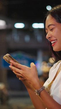Uzun düz saçlı Asyalı bir kız, Bangkok 'taki FullHD Dikey Caddesi' nde gece vakti akıllı telefonuyla bir video görüşmesi yapıyor.