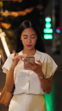 Asyalı genç bir kadın sıcak bir gecede akıllı telefon ekranına dokunarak mutlu bir şekilde büyük caddede yürüyor.