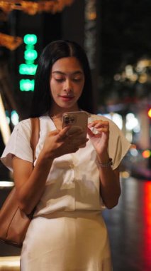 Asyalı genç bir kadın gece büyük caddede dikilirken akıllı telefonuyla mesajlaşırken gülümsüyor.
