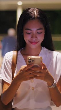 Genç Asyalı bir kadın geceleri FullHD dikey kaldırımda yürürken akıllı telefon ekranına dokunuyor.