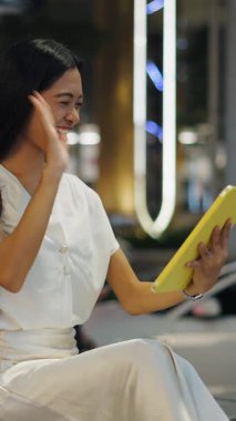 Genç Asyalı bir kadın büyük caddenin yanında kaldırımda oturmuş elinde tabletle video görüşmesi yapıyor.