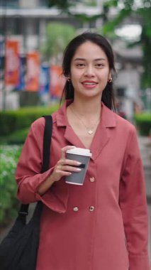 Asyalı genç bir iş kadını elinde kahve tutarak şehir caddesinde yürüyor.