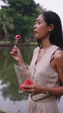 Genç bir kadın, sakin bir göl kenarında taze bir karpuz diliminin tadını çıkarıyor - FullHD Dikey