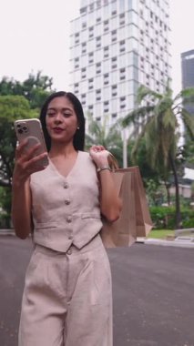 Sokakta akıllı telefonuyla yürüyen ve alışveriş torbaları taşıyan genç bir kadın.