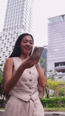 Şehir parkında akıllı telefonuyla yürüyen ve gülümseyen genç bir iş kadını.