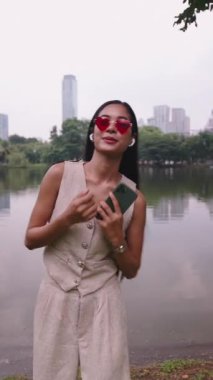 Kalp şeklinde güneş gözlüğü takan genç bir kadın parkta akıllı telefon kullanıyor.