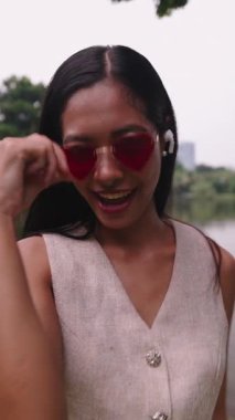 Şık bir kadın, sakin bir göl parkında müzik dinlerken kalp şeklindeki güneş gözlüğünü ayarlar.