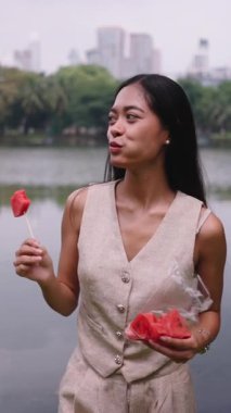 Güzel genç bir kadın şehir parkındaki bir gölün yanında taze karpuz yiyor.