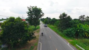 Tayland kırsalındaki yemyeşil bitki örtüsüyle çevrili asfalt bir yolda seyahat eden araçlar - 4K Yatay