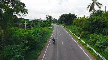 Etrafı yemyeşil bitkilerle çevrili kırsal bir yolda seyahat eden motosikletçi - 4K Yatay