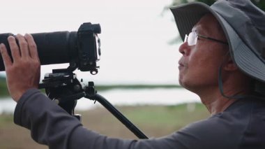 Asyalı fotoğrafçının profili, büyük şapkalı, tripodu ayarlayan ve refleks kameranın yakınlaştırdığı - 4K Yatay