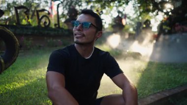 Bahçede oturan kameraya gülümseyen büyük gözlüklü genç bir Latin 'in büstü. 4K Yatay