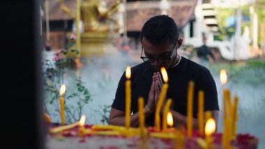 Genç Latin çocuk Budist tapınağının önünde diz çöküp mum yakarak dua ediyor. 4K Yatay