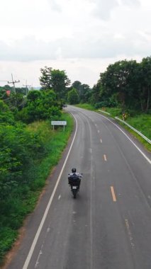 Çevresi yemyeşil bitkilerle çevrili kırsal bir yolda seyahat eden motosikletçi FullHD Dikey