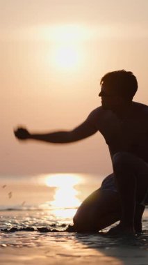 Gün batımında kumsala ıslak kum atan genç bir adamın silueti FullHD Dikey