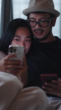 Genç ve mutlu Taylandlı çift, akıllı telefonlarını kullanarak kanepede dinleniyorlar.