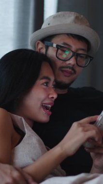 Mutlu Asyalı çift rahat bir kanepede rahat rahat oturup akıllı telefondan sosyal medyaya bakıyorlar.