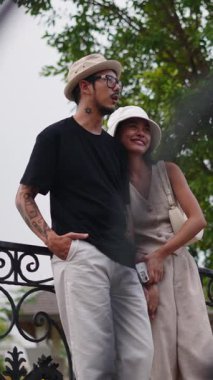 Genç Taylandlı çift parkta romantik bir anın tadını çıkarıyor korkuluklara yaslanıyor ve gülümsüyor.