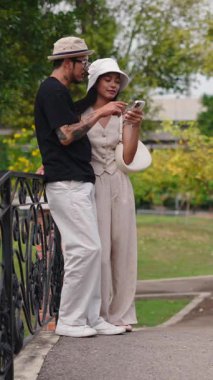 Genç Çok Irklı Çift, Parktaki Köprü korkuluklarına yaslanırken Akıllı Telefon Kullanıyor