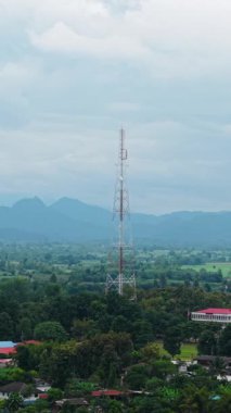 Bitkilerle çevrili bir köyün üzerinde yükselen telekomünikasyon kulesinin resimli görüntüsü - FullHD Dikey