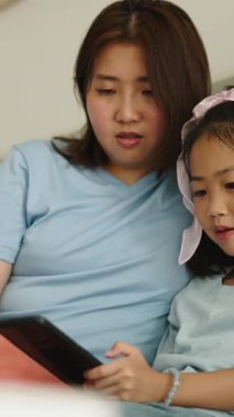 Asyalı genç anne ve kız kanepe üzerinde dijital tablet paylaşıyorlar.