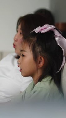 Asyalı anne ve kızı oturma odasında televizyon seyrediyor ve ekrandaki bir şeyi işaret ediyor.