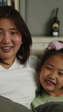 Asyalı anne ve kızı kanepede gülüyor sessiz bir oturma odasında sarılıyorlar - FullHD Dikey