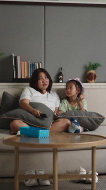 Asyalı anne ve kız bir kanepede oturup televizyon seyrediyor ve rahatlatıcı bir anı paylaşıyorlar.