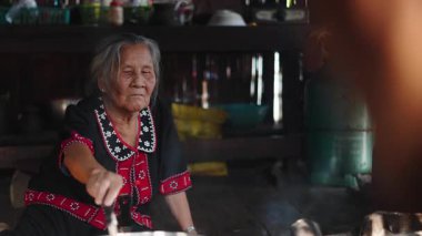 Geleneksel kıyafetler giymiş yaşlı bir kadın, kırsal bir mutfakta sıcak bir tencerede yemek hazırlıyor - 4K Yatay