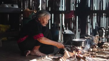 Geleneksel lahu kıyafetleri giymiş yaşlı bir kadın kulübede odun ateşinde yemek hazırlıyor - 4K Yatay
