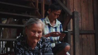 Yaşlı bir kadın tahta merdivenlerde yanında otururken yaşlı bir adam akıllı telefon kullanıyor. 4K Yatay